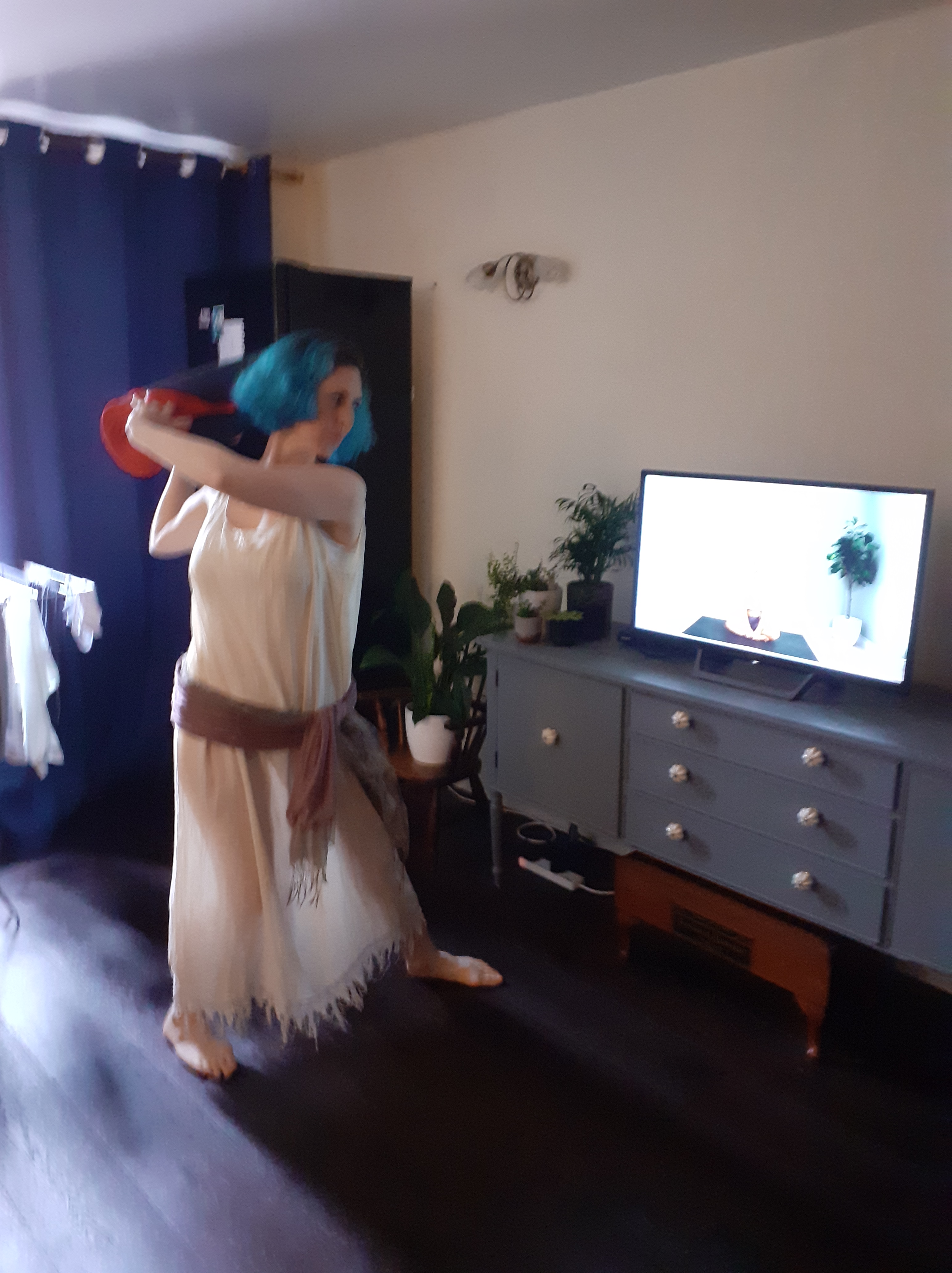 Georgi wearing a Grecian dress is standing in a sitting room. She is holding a yoga mat and poised to hit a television with the mat.
